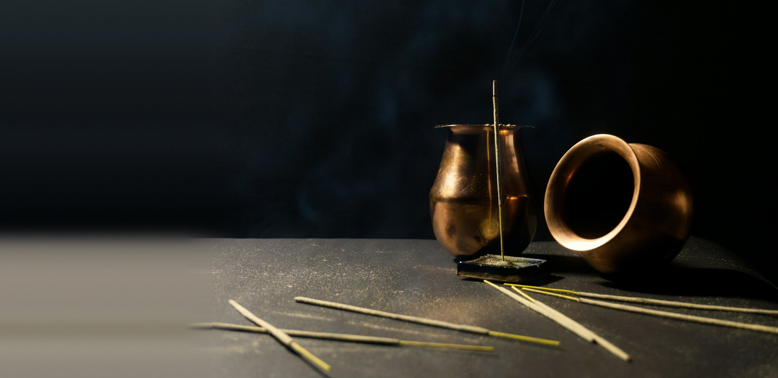 A lit incense stick standing between two copper vessels, with loose sticks on a dark table